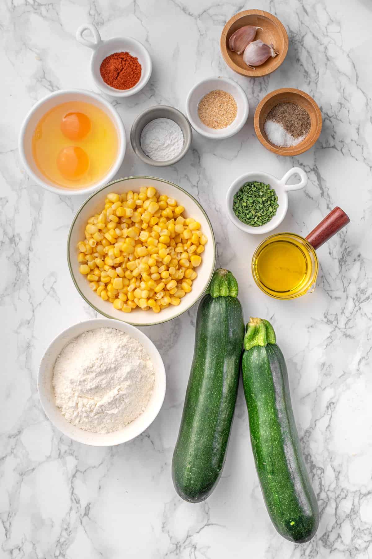 Zucchini and Corn Fritters ingredients on marble: 2 zucchinis, corn kernels, eggs, all-purpose flour, olive oil, smoked paprika, garlic cloves, dried spices