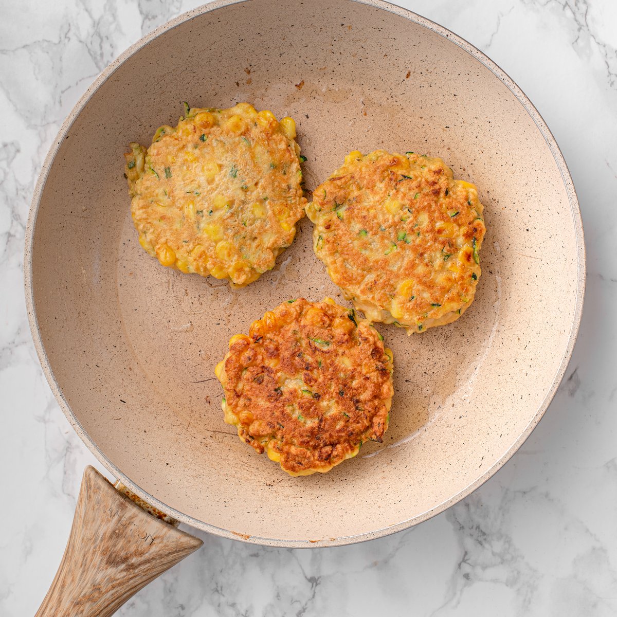 Three zucchini and corn fritters cooking in a beige nonstick skillet, two golden-brown and one lighter side up, wooden spatula visible.