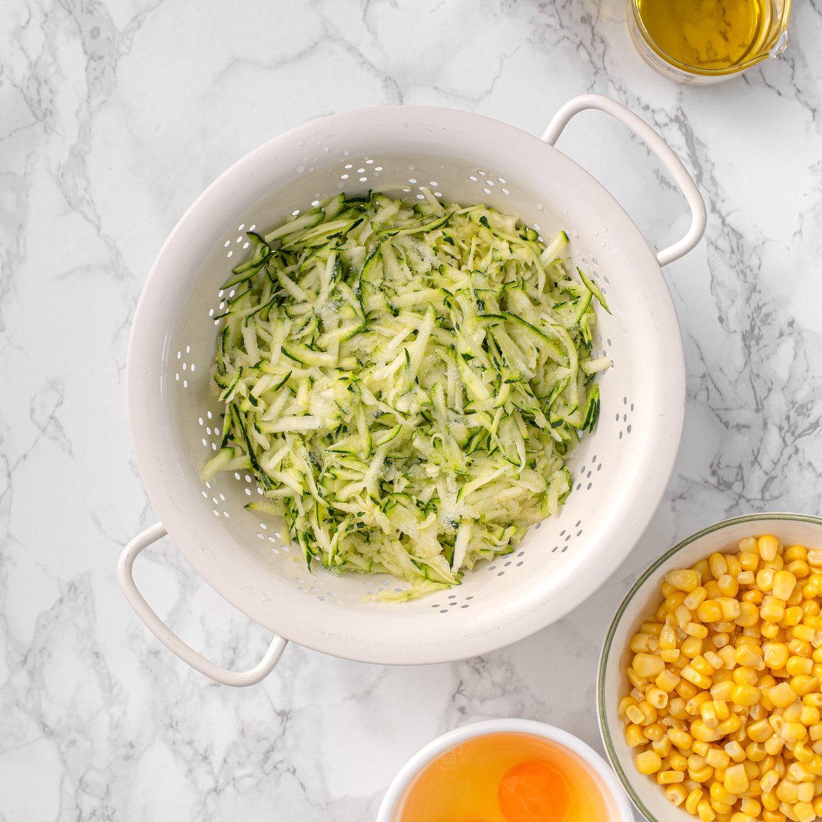 Grated zucchini draining in a white colander beside bowls of corn kernels, an egg, and olive oil on a marble surface.