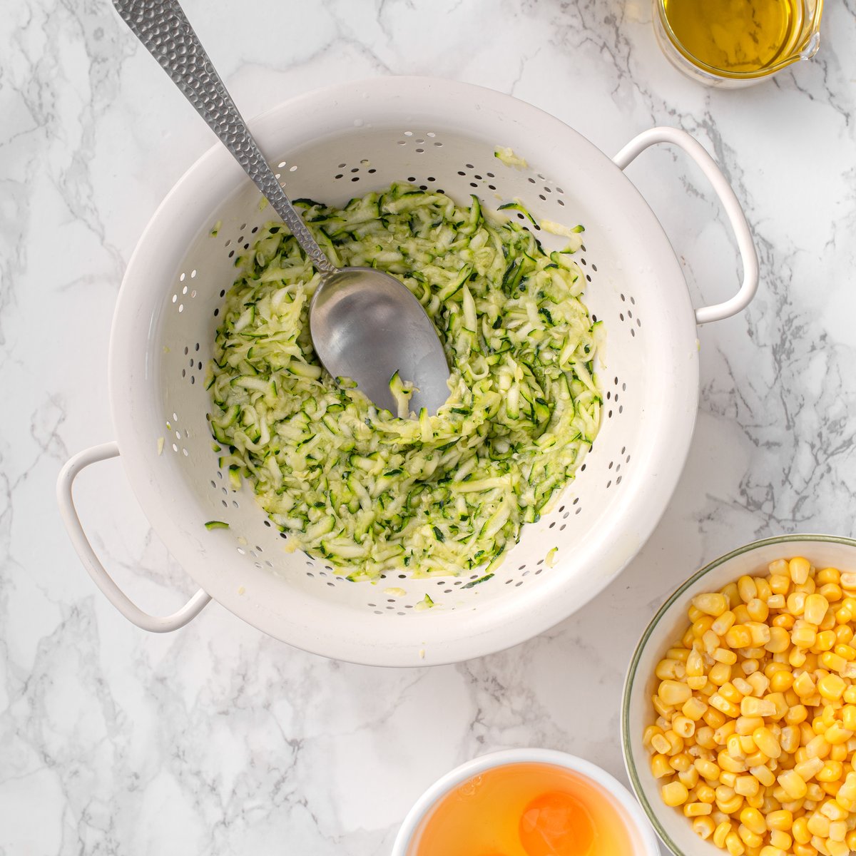 Grated zucchini draining in a white colander with a spoon, beside bowls of corn kernels and an egg on a marble surface.