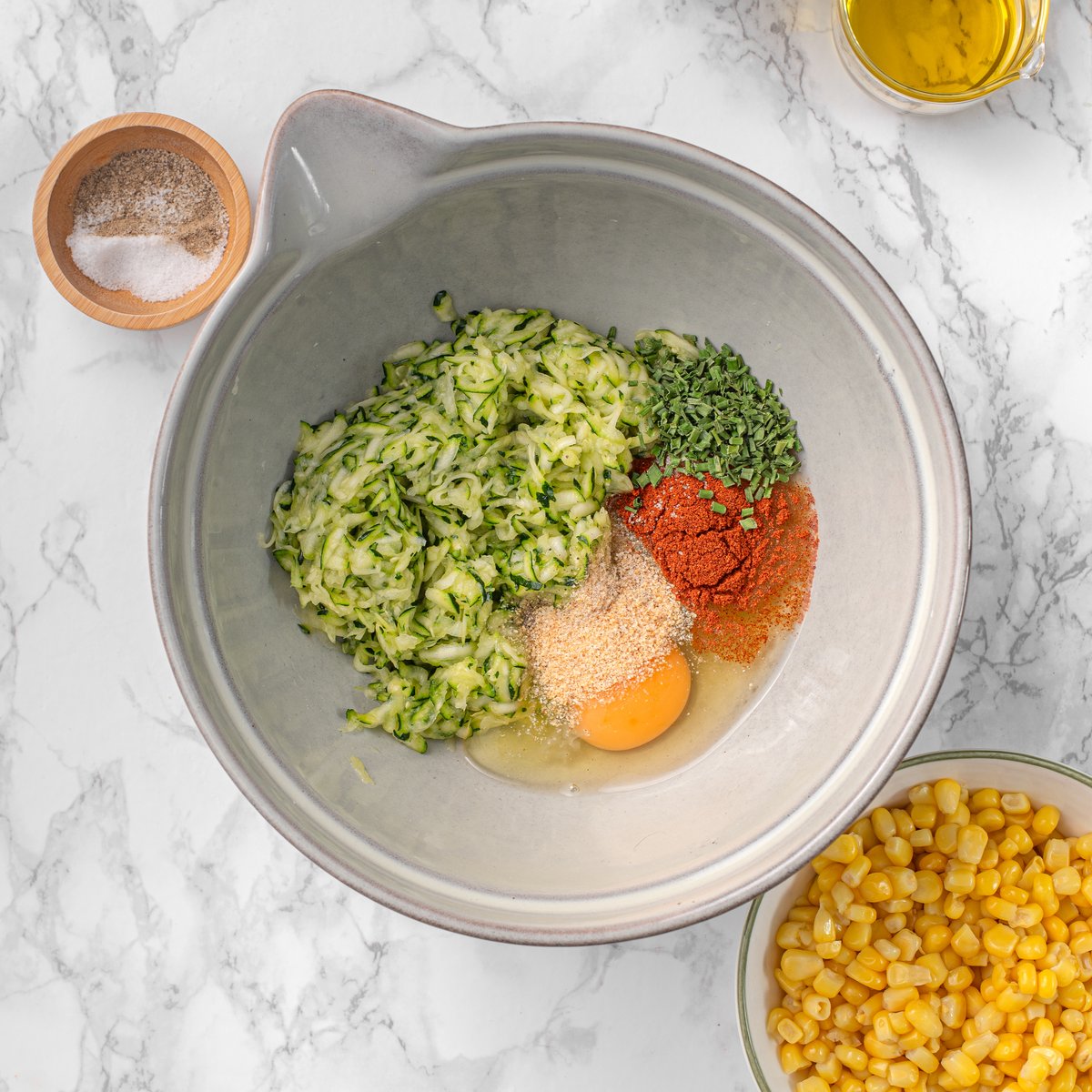 Mixing bowl with grated zucchini, egg, smoked paprika, onion powder, and dried chives for zucchini and corn fritters