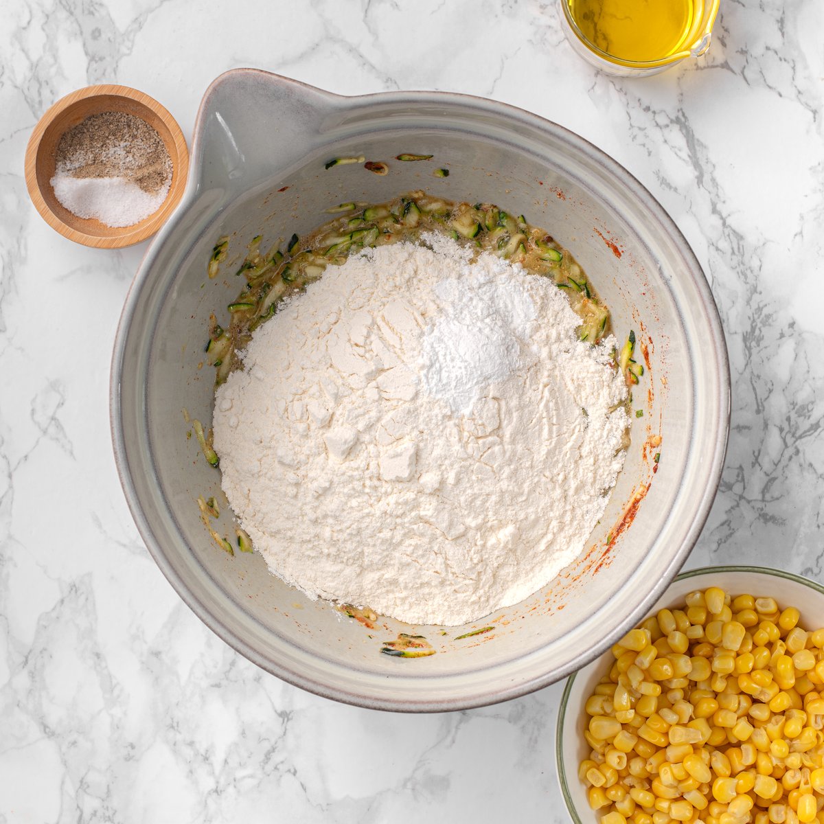 Mixing bowl with all-purpose flour added over grated zucchini batter, with corn kernels, olive oil, and salt and black pepper nearby on marble.