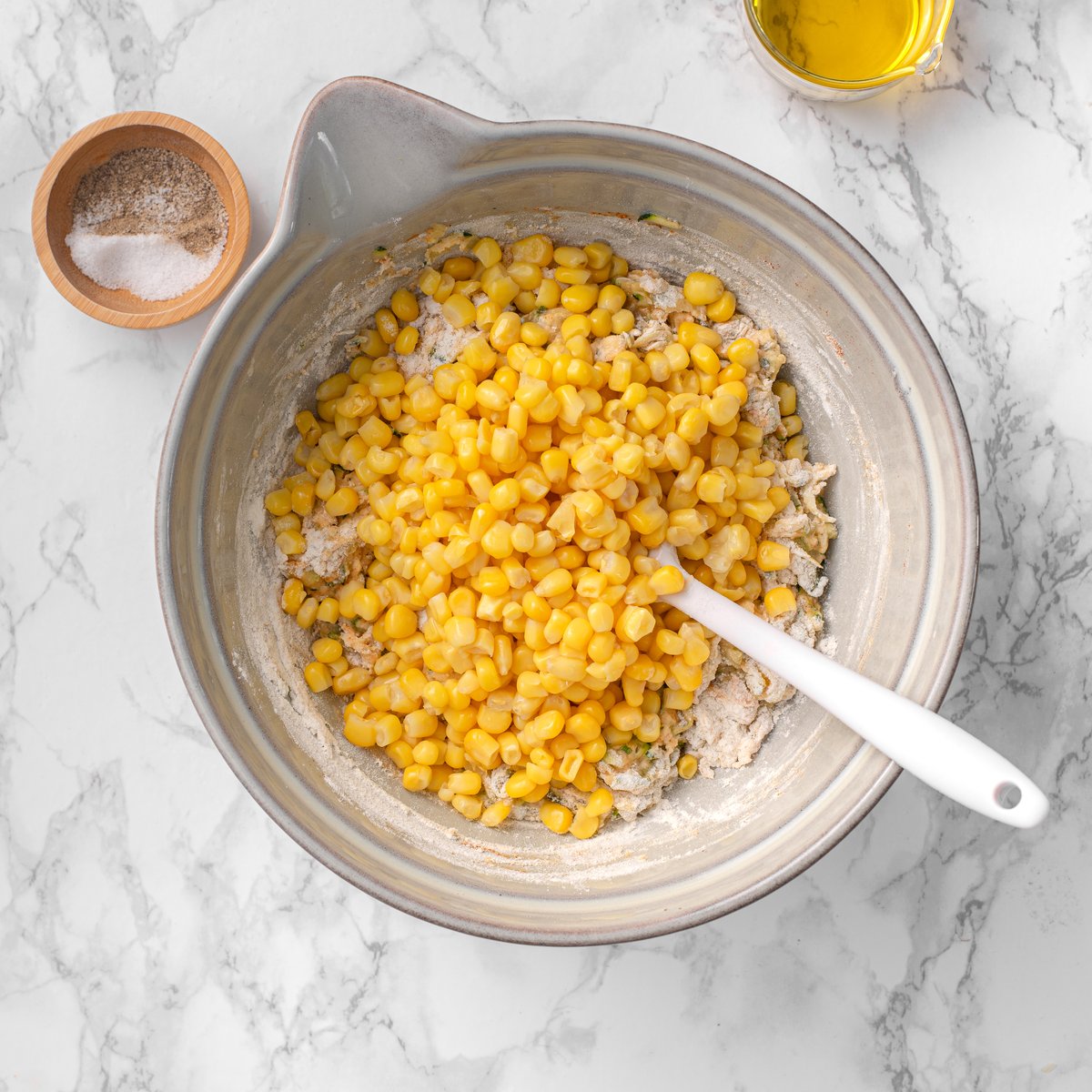 Corn kernels added to zucchini fritter batter in a gray bowl with a white spatula on a marble surface.