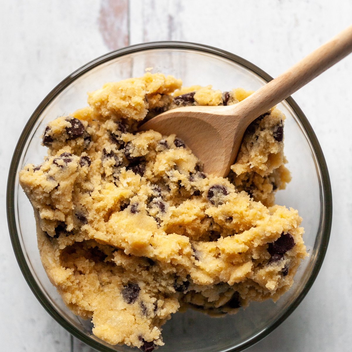 Chickpea cookie dough with dairy-free chocolate chips in a glass bowl being stirred with a wooden spoon on a white surface.