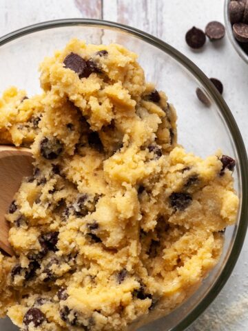 Chickpea cookie dough mixed in a glass bowl with a wooden spoon, studded with dairy-free chocolate chips, with extra chips in a small bowl nearby.