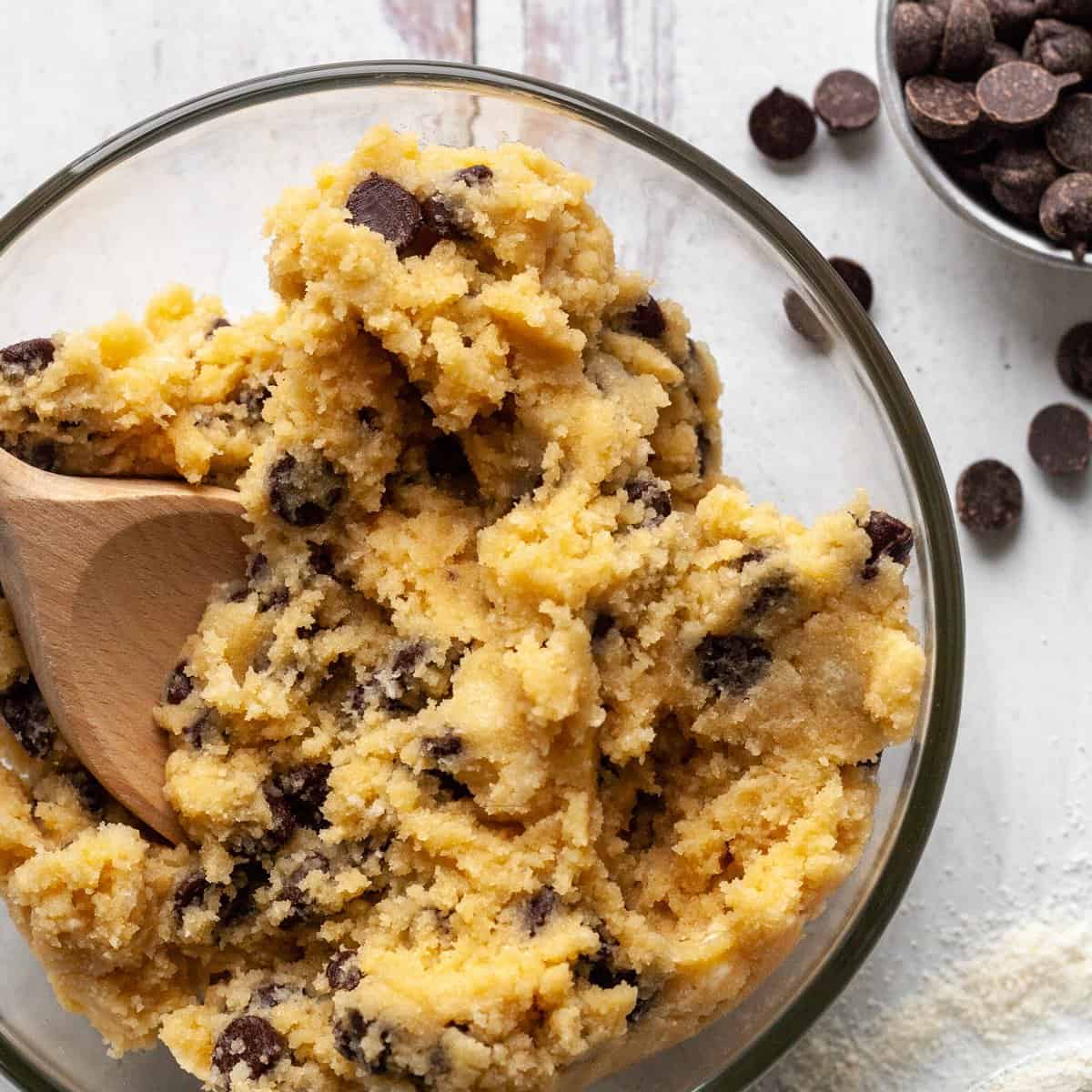 Chickpea cookie dough mixed in a glass bowl with a wooden spoon, studded with dairy-free chocolate chips, with extra chips in a small bowl nearby.