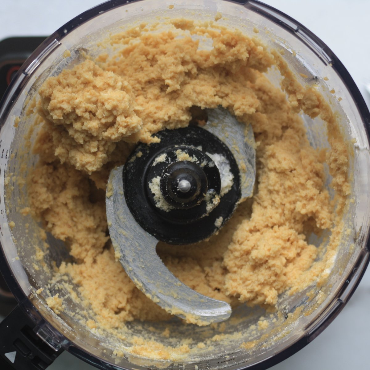 Chickpea cookie dough blended in a food processor bowl showing thick, crumbly pale golden mixture around the blade.