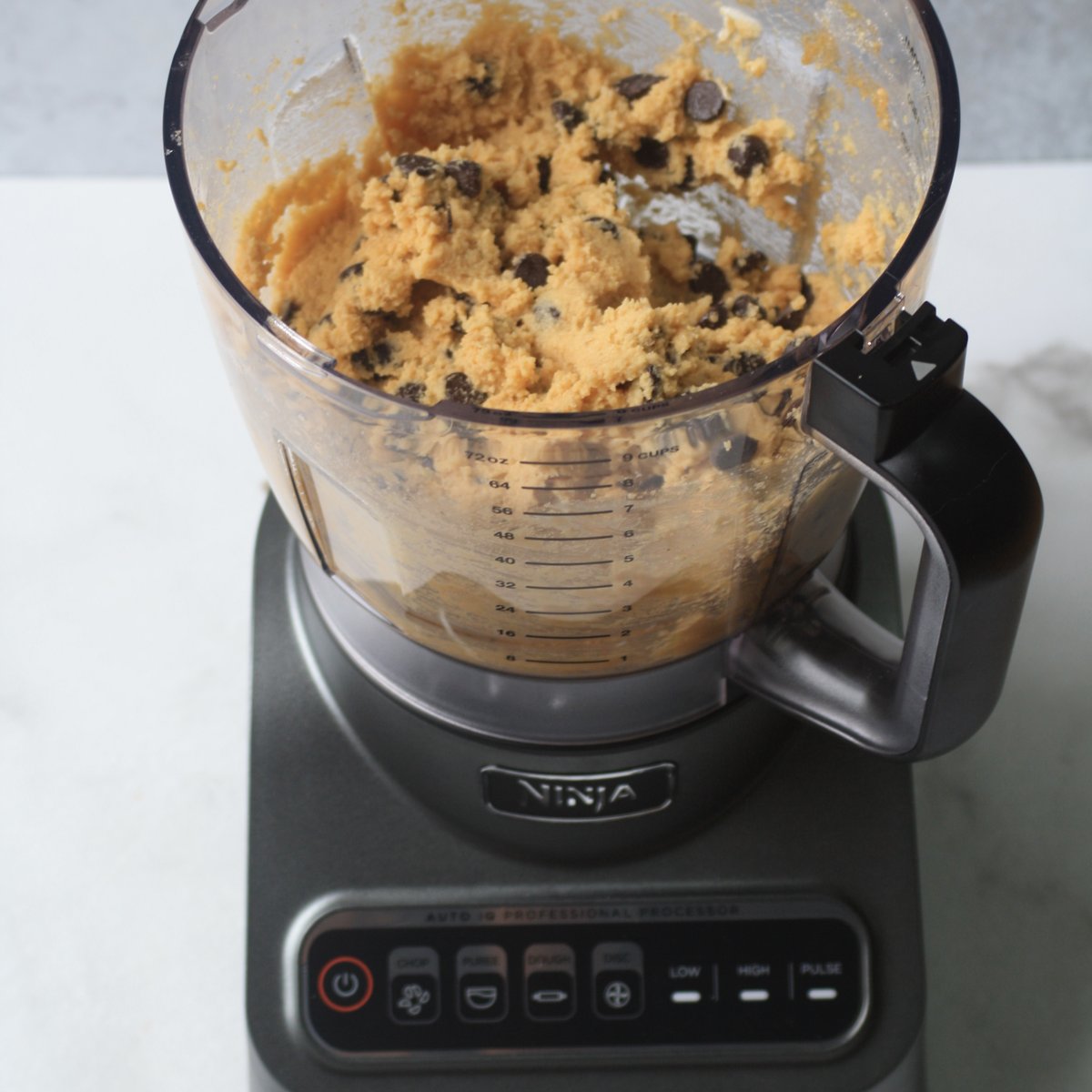 Ninja food processor bowl filled with blended chickpea cookie dough and dairy-free chocolate chips on a marble surface
