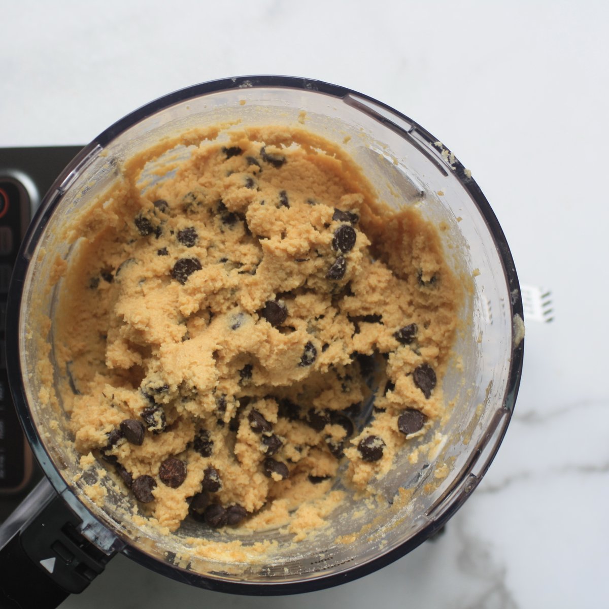 Chickpea cookie dough with dairy-free chocolate chips mixed in a food processor bowl on a marble surface.