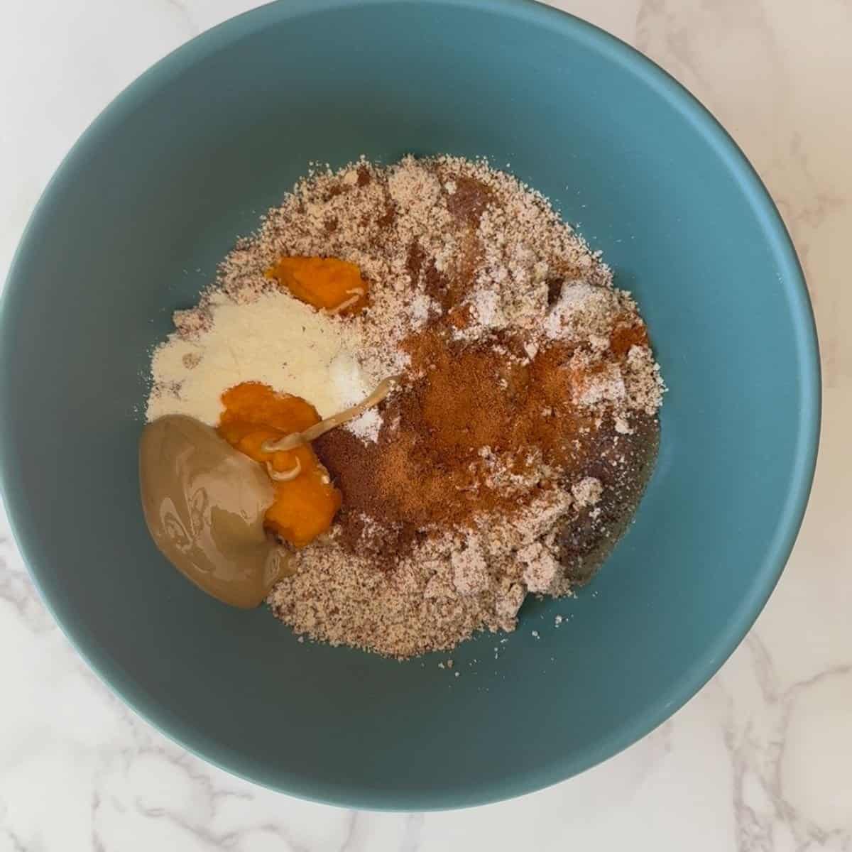 Teal bowl with almond flour, vanilla protein powder, pumpkin puree, sunflower butter, and allspice powder for pumpkin spice protein balls