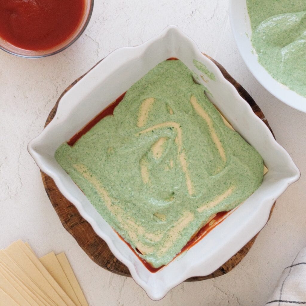 Spinach ricotta filling spread over noodles in a square white baking dish, with marinara sauce and lasagna noodles alongside.