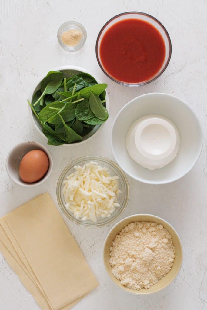 Spinach ricotta lasagna ingredients: fresh spinach, marinara sauce, whole milk ricotta, shredded mozzarella, grated Parmesan, egg, seasonings, and lasagna noodles.