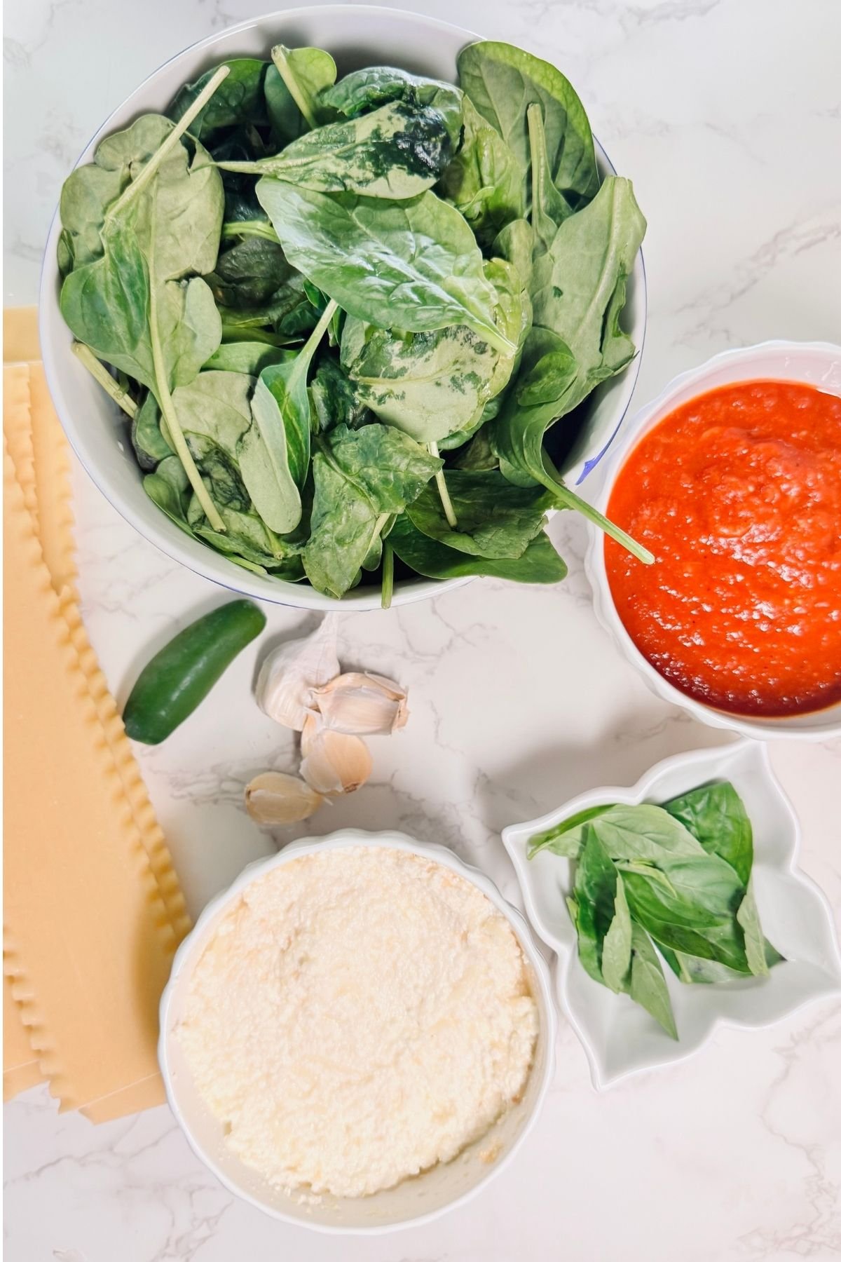 Spinach Ricotta Lasagna ingredients on marble: fresh spinach, whole milk ricotta, marinara sauce, garlic cloves, lasagna noodles, and fresh basil