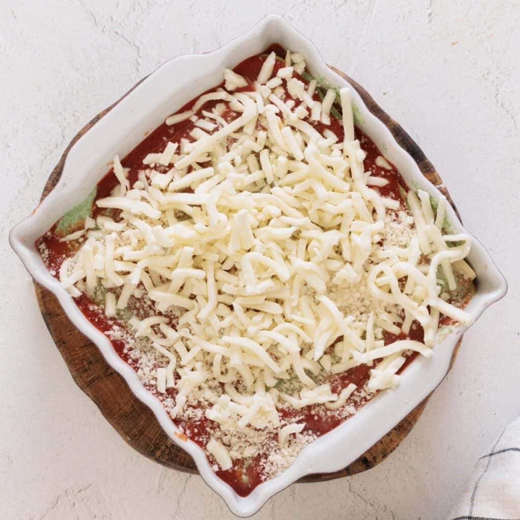 Assembled spinach ricotta lasagna in a square baking dish topped with marinara, shredded mozzarella, and Parmesan, ready to bake.