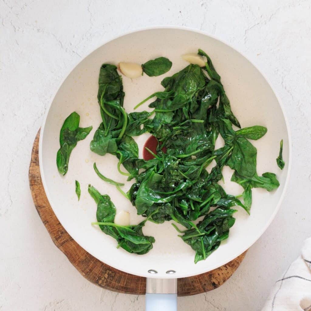 Fresh spinach and whole garlic cloves sauteed in a white skillet for spinach ricotta lasagna filling.