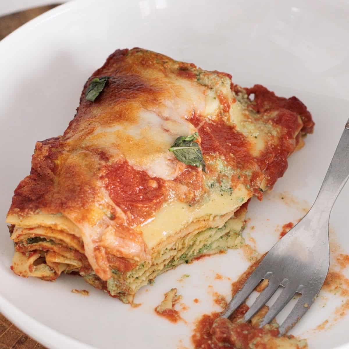 Close-up of a spinach ricotta lasagna slice in a white bowl with a fork, showing layered noodles and spinach ricotta filling.