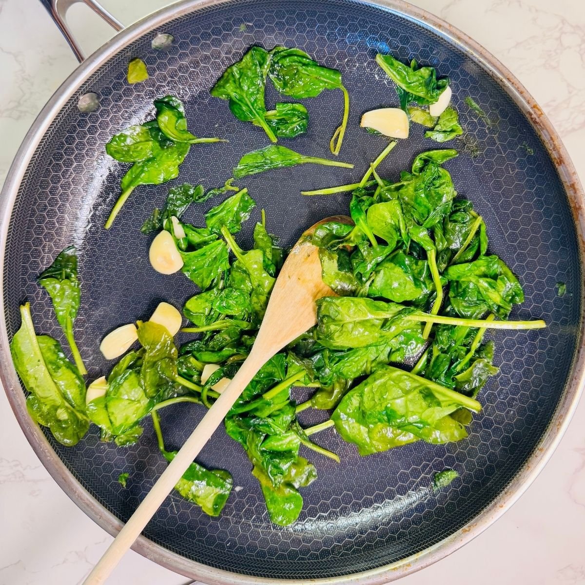 Fresh spinach sautéed with peeled garlic cloves in a skillet with a wooden spoon for spinach ricotta lasagna filling.