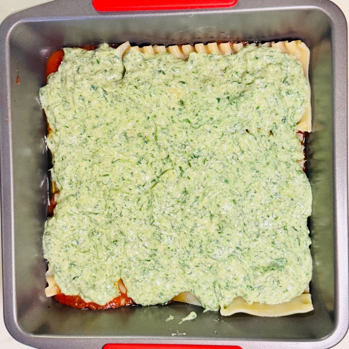 Spinach ricotta lasagna being assembled in a square baking pan with spinach ricotta mix spread over lasagna noodles and marinara sauce layer.