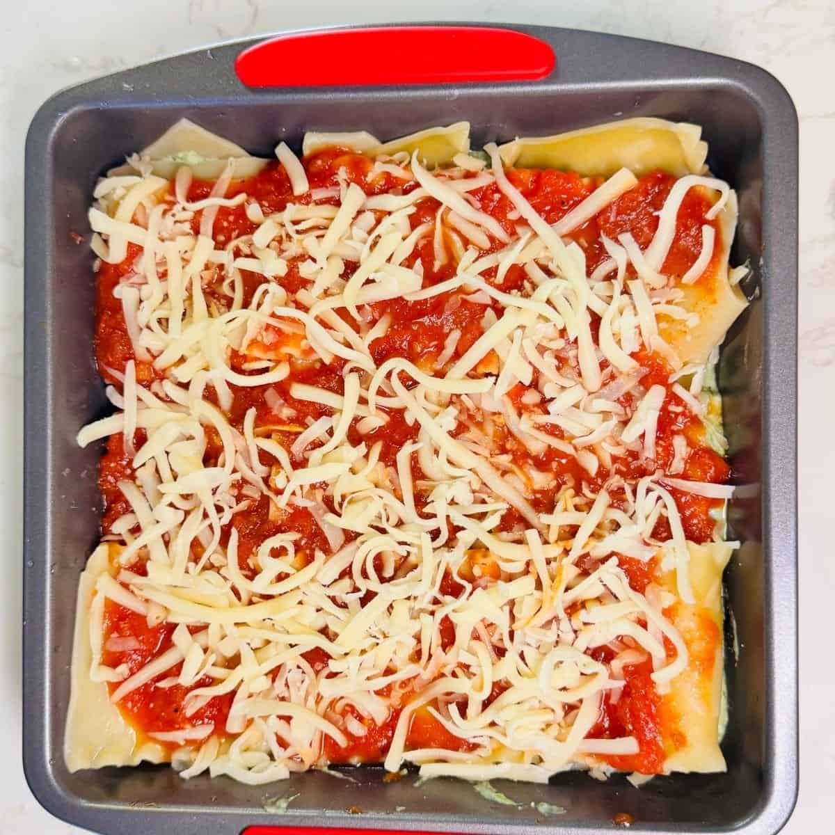 Unbaked spinach ricotta lasagna in a square pan topped with marinara sauce and shredded mozzarella ready to bake.