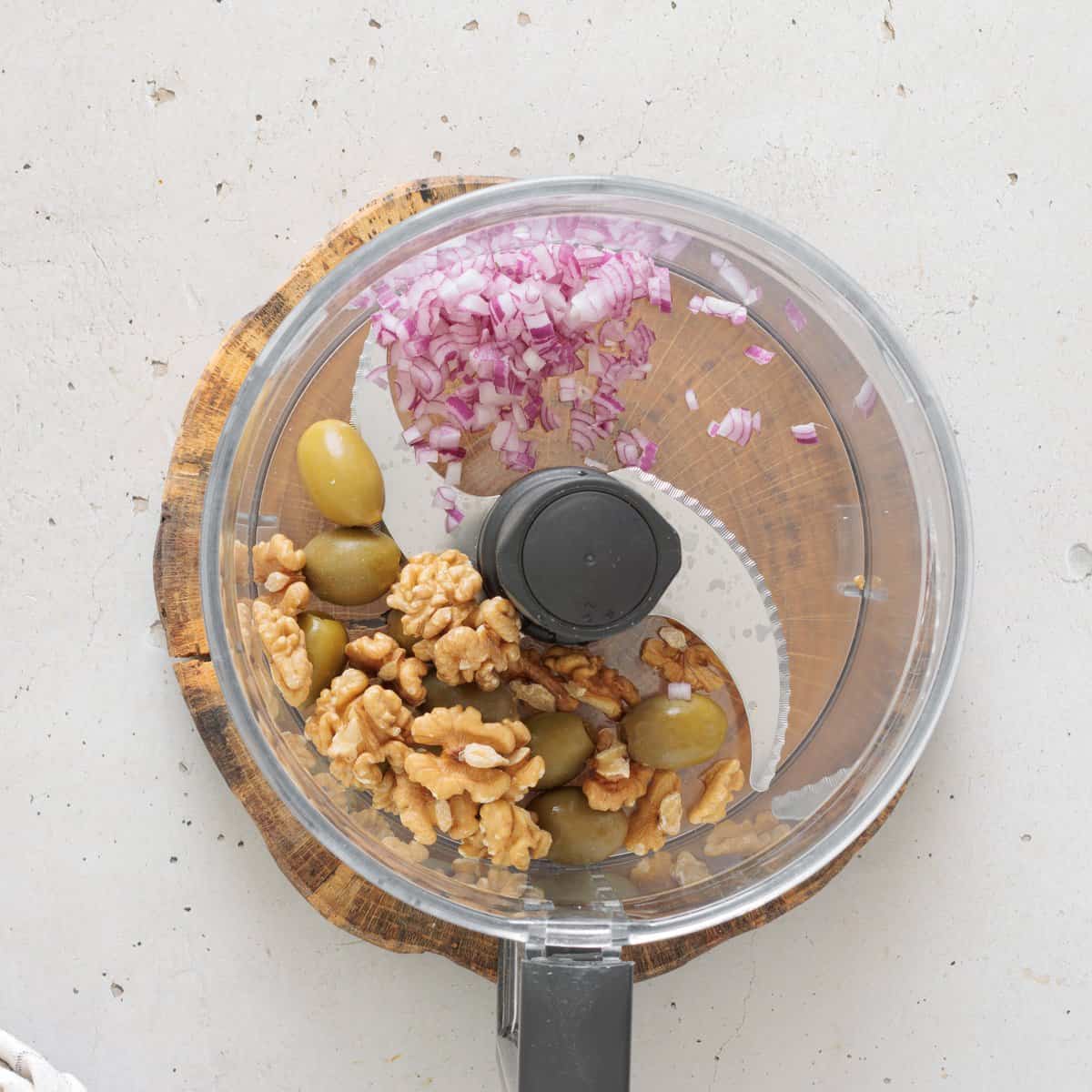 Food processor bowl with diced red onion, walnuts, and olives for chickpea salad on a wooden board