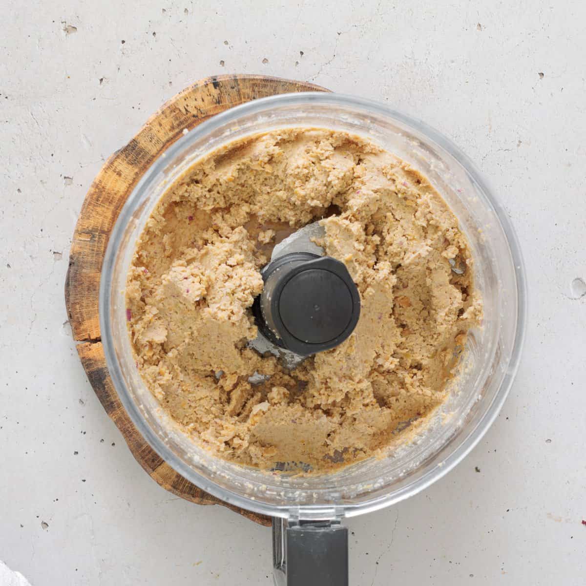Chickpea salad mixture with olives and walnuts blended into a coarse paste inside a food processor bowl on a wooden board.