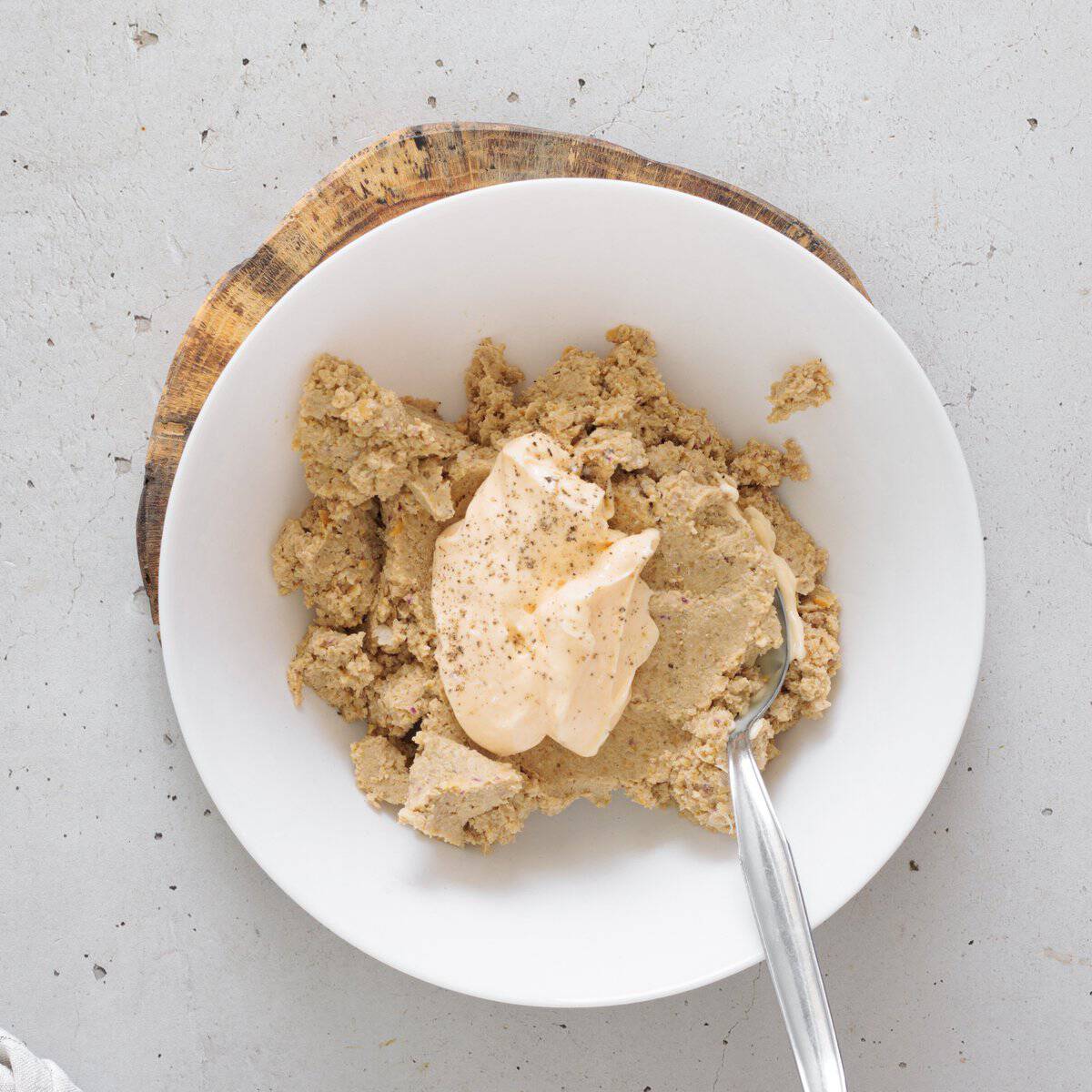 Mashed chickpeas with a dollop of creamy mayo and black pepper in a white bowl with a spoon on a wooden board