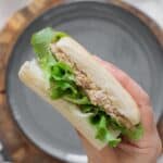 Hand holding a chickpea salad sandwich with olives, walnuts, and lettuce on a white roll over a gray plate on a wood board.