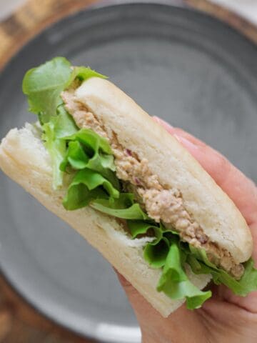 Hand holding a chickpea salad sandwich with olives, walnuts, and lettuce on a white roll over a gray plate on a wood board.