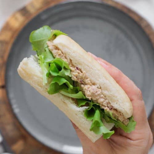 Hand holding a chickpea salad sandwich with olives, walnuts, and lettuce on a white roll over a gray plate on a wood board.