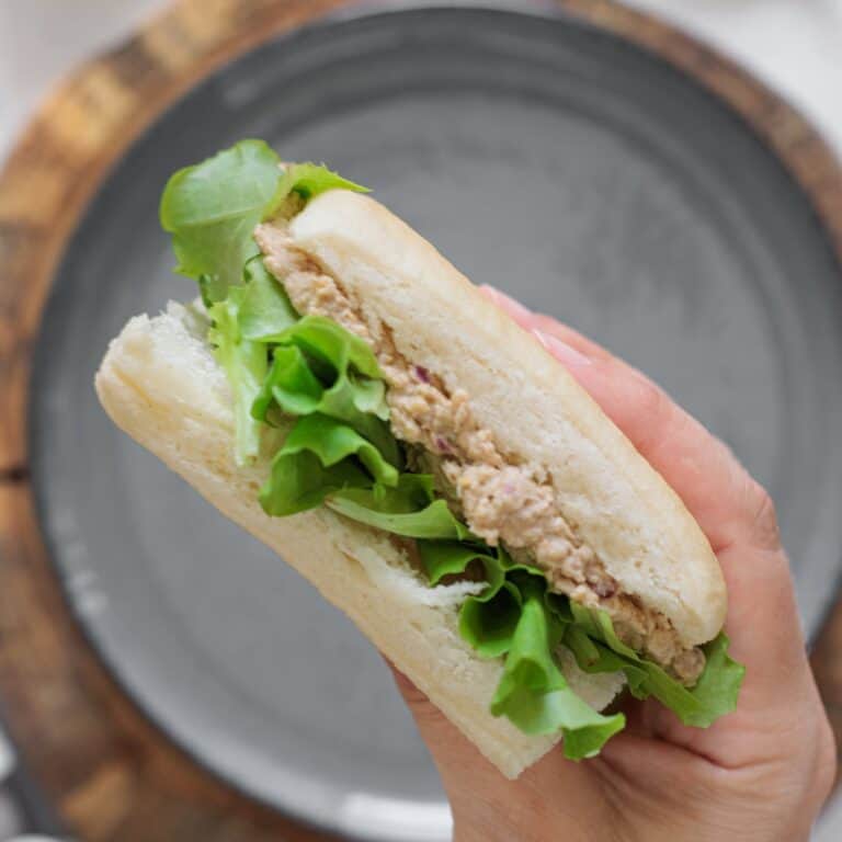 Hand holding a chickpea salad sandwich with olives, walnuts, and lettuce on a white roll over a gray plate on a wood board.