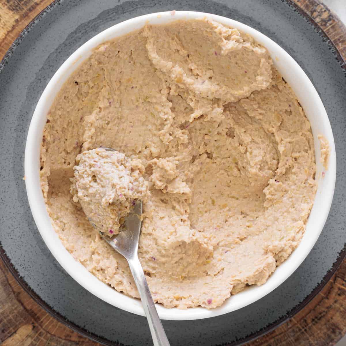 Creamy chickpea salad with olives and walnuts in a white bowl with a spoon, served on a gray plate over a wood surface
