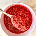 Raspberry chia jam in a white bowl with a spoon lifting a scoop, showing glossy red texture with chia seeds on marble