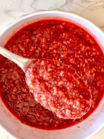 Raspberry chia jam in a white bowl with a spoon lifting a scoop, showing glossy red texture with chia seeds on marble