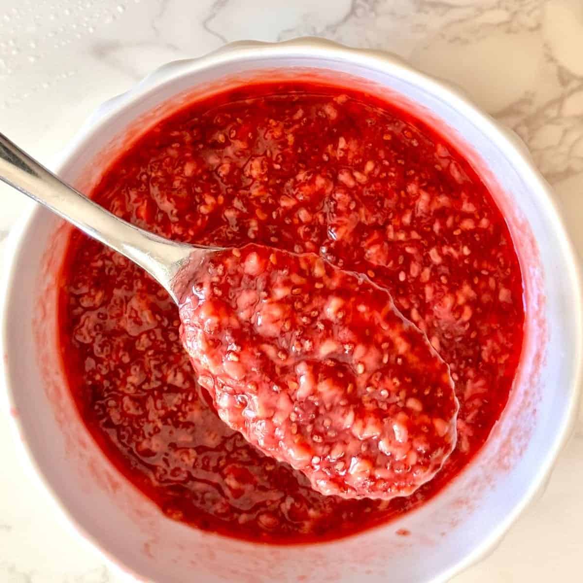 Raspberry chia jam in a white bowl with a spoon lifting a scoop, showing glossy red texture with chia seeds on marble