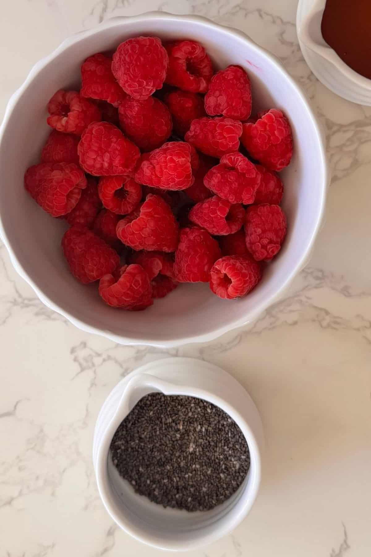 Fresh raspberries in a white bowl beside a small white dish of chia seeds for Raspberry Chia Jam on a marble surface