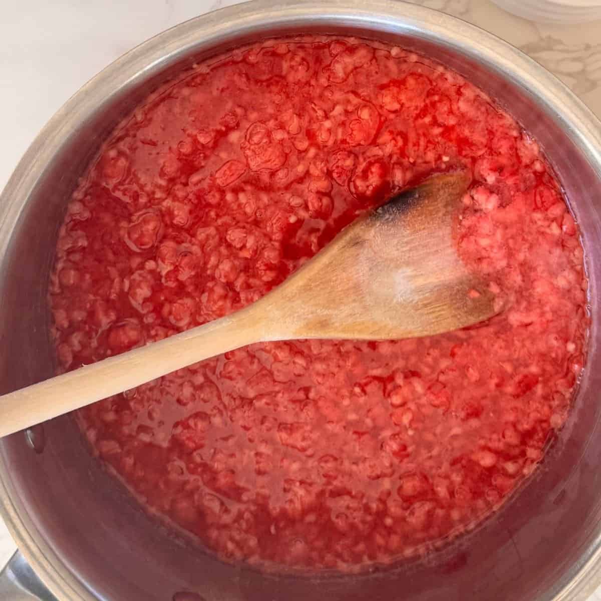 Fresh raspberries simmering into Raspberry Chia Jam in a steel pot, stirred with a wooden spoon