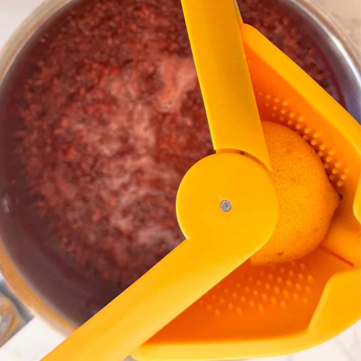 Squeezing ¼ lemon with a yellow citrus press over a pot of simmering fresh raspberries for raspberry chia jam