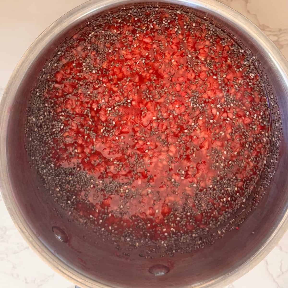 Raspberry chia jam bubbling in a stainless steel saucepan with mashed fresh raspberries and chia seeds on a marble surface.